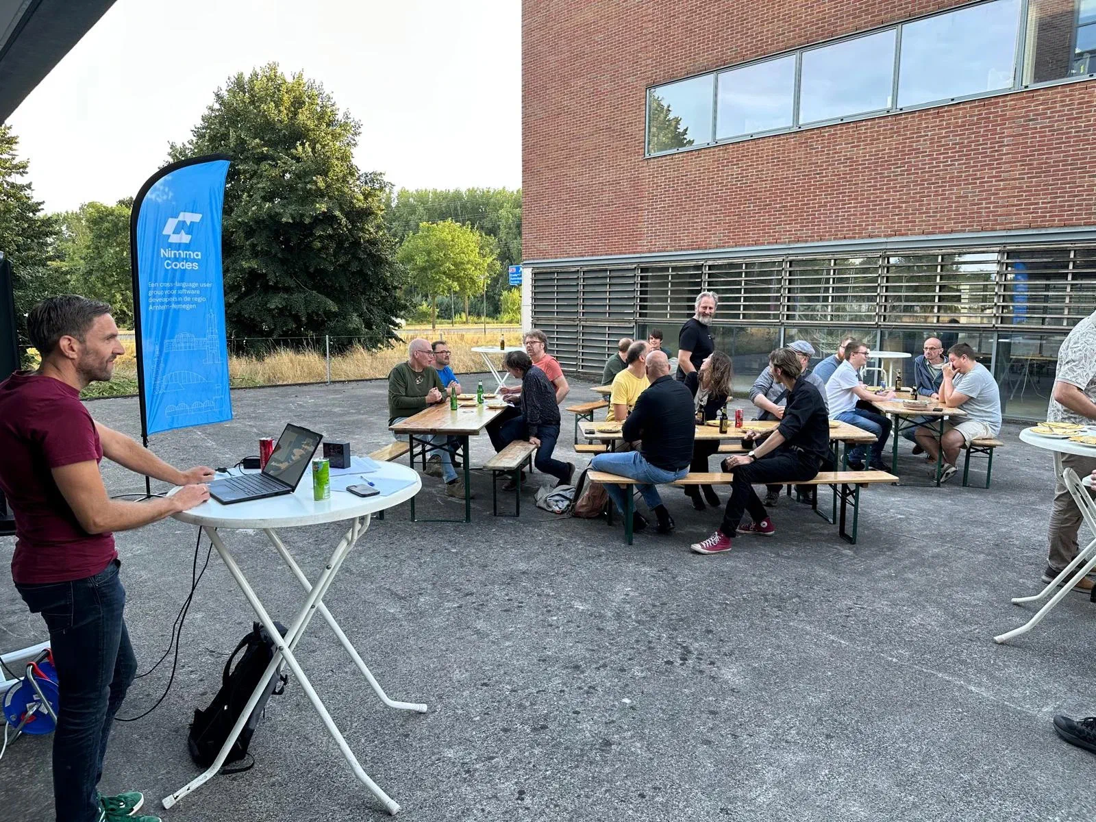 Bezoekers zitten buiten aan picknicktafels terwijl iemand bij een statafel met laptop praat